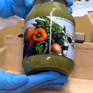 A glass jar filled with fresh, green Haitian Epis seasoning, surrounded by fresh garlic cloves, green onions, and red bell peppers.